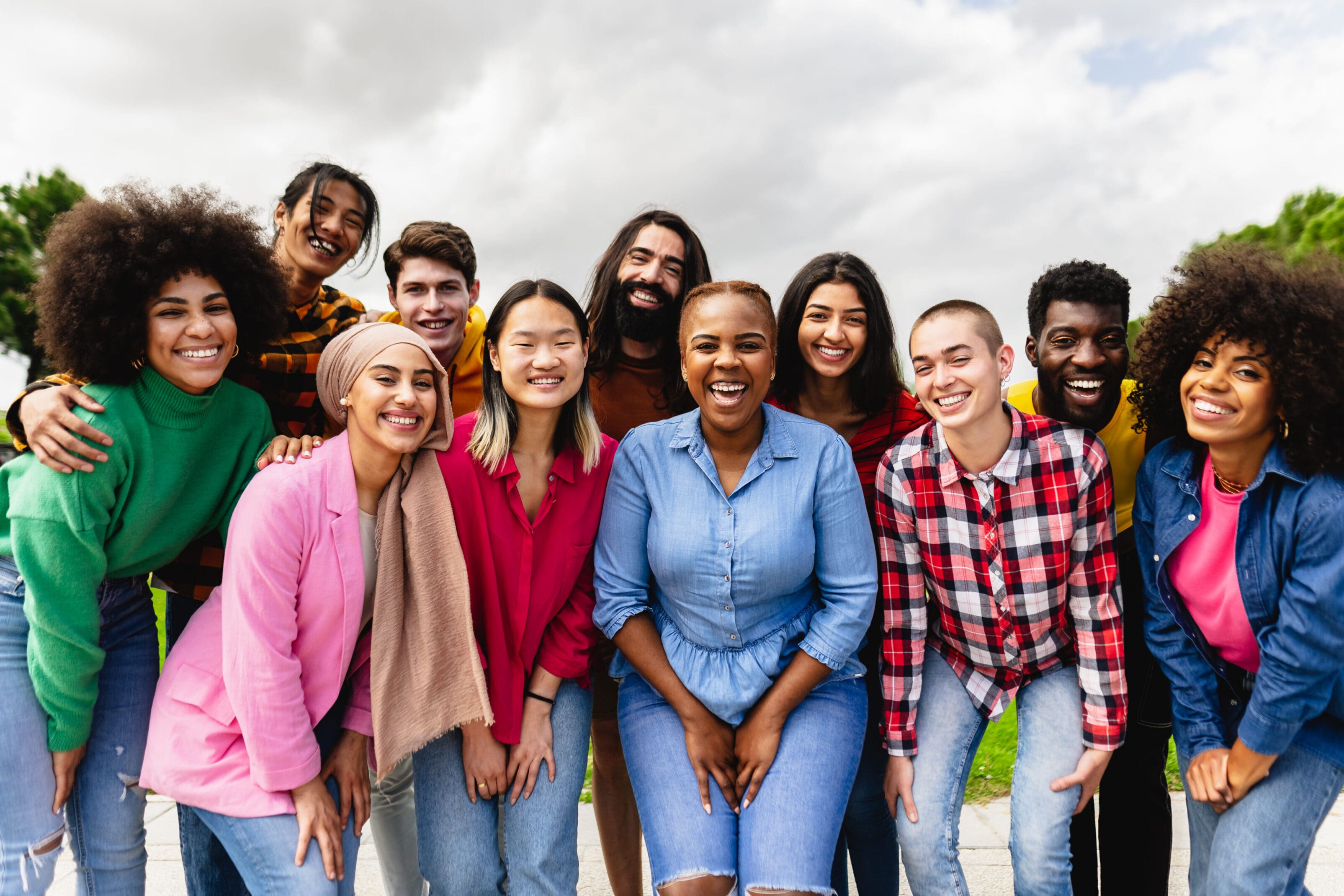 A group of thirteen diverse young adults stand close together outdoors, smiling at the camera under a cloudy sky.