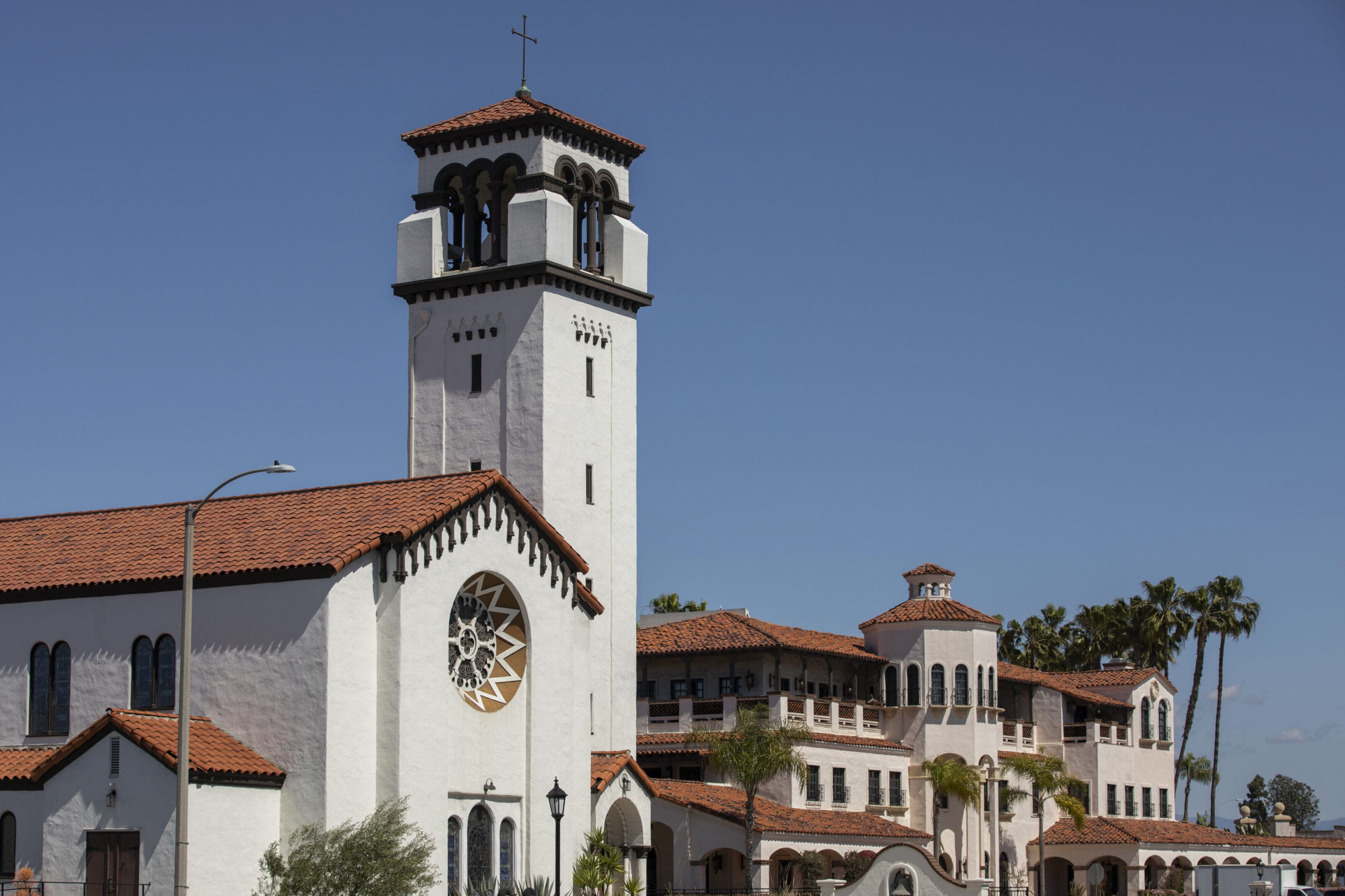A large white stucco church with a red tile roof, a tall bell tower topped with a cross, and a circular stained-glass window, set against a clear blue sky.