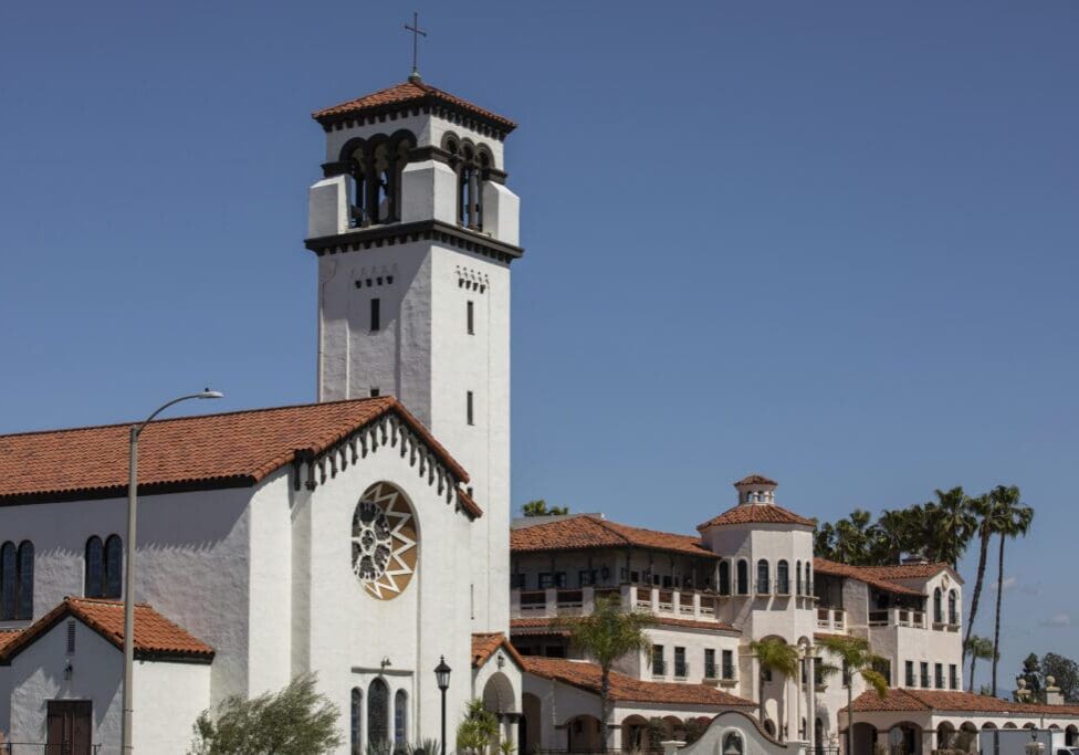 A large white stucco church with a red tile roof, a tall bell tower topped with a cross, and a circular stained-glass window, set against a clear blue sky.